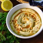 Hummus with tahini in a white bowl, drizzeled with olive oil and pine nuts.