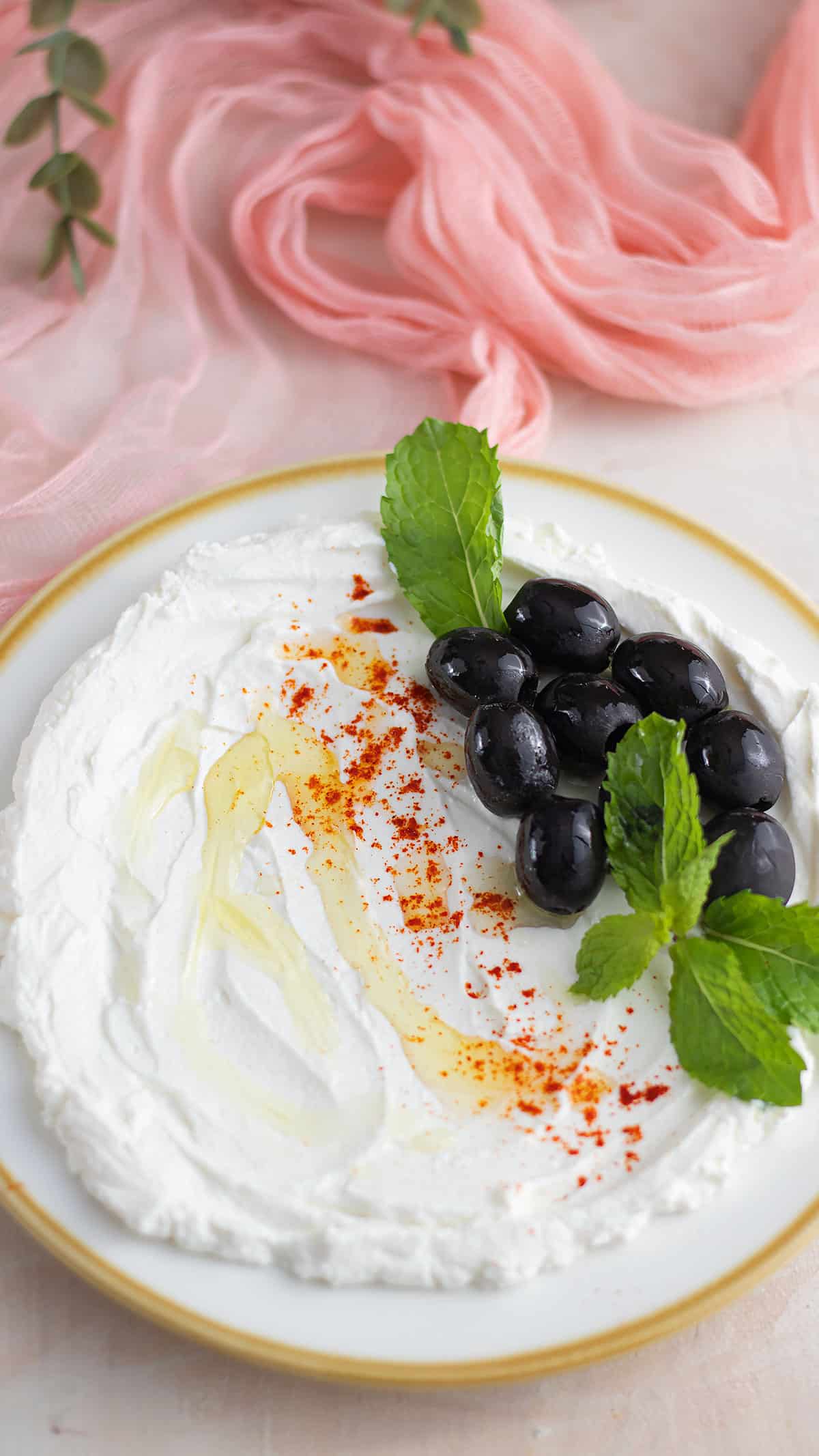 Homemade labneh spread evenly on a white plate, drizzeled with olive oil, and garnished with black olives on the side with a few mint leaves. 1