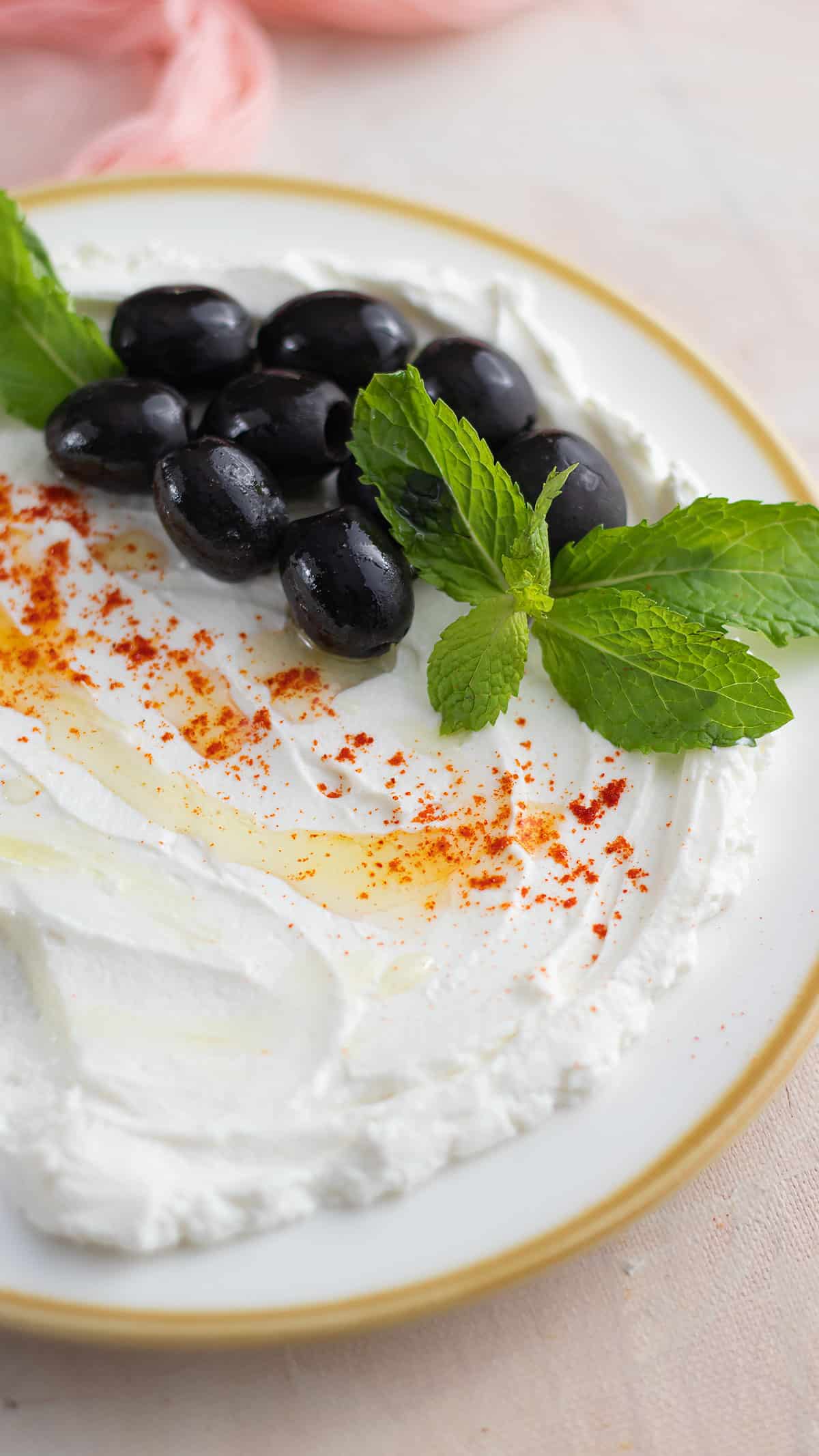 Close up image of labneh spread on a white plate garnished with black olives, mint leaves and drizzele of olive oil.